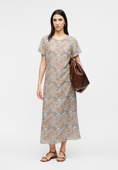 Γυναίκα με ίσια σκούρα μαλλιά, που φοράει μπλε και μπεζ midi φόρεμα με σχέδιο, ταμπά σανδάλια, χρυσούς κρίκους στα αυτιά και κρατάει μια μεγάλη καφέ δερμάτινη τσάντα.