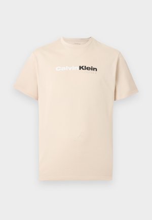 Μπεζ μπλουζάκι με κοντά μανίκια και στρογγυλή λαιμόκοψη, με το λογότυπο "Calvin Klein" σε λευκό και μαύρο στο κέντρο του στήθους.