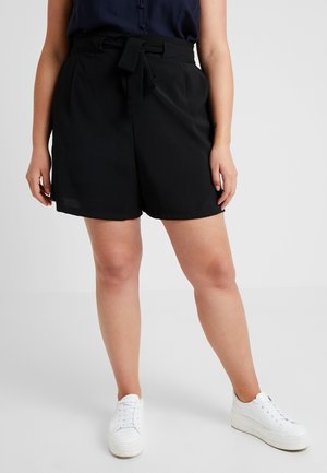 Femme portant un short noir taille haute à nouer devant, un chemisier bleu marine et des baskets à plateforme blanches, debout sur un sol et un fond gris clair.