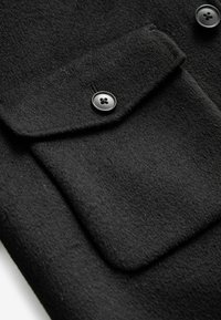 Detaliu de țesătură din lână neagră cu o buzunar cu clapetă, având un buton rotund negru cu un design cu patru găuri. Textură moale și finisare netedă.