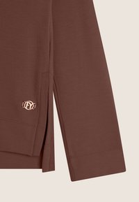 Top marrone a maniche lunghe con fessure laterali, texture liscia e un piccolo logo metallico accentato in basso a sinistra.