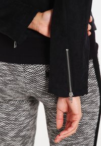 Veste en daim noir avec des détails de poignet zippés. Le porteur porte un pantalon à motifs noir et blanc, soulignant le contraste de textures.