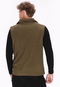 Olivgrüne Fleeceweste mit hohem Kragen und ohne Ärmel, glatte Textur und gerader Saum. Getragen über einem schwarzen Langarmshirt.