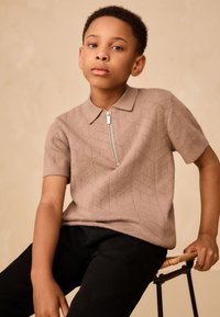 Tricou polo bej tricotat cu model geometric, mâneci scurte și închidere cu fermoar argintiu. Purtat cu pantaloni negri, așezat pe un scaun înalt.