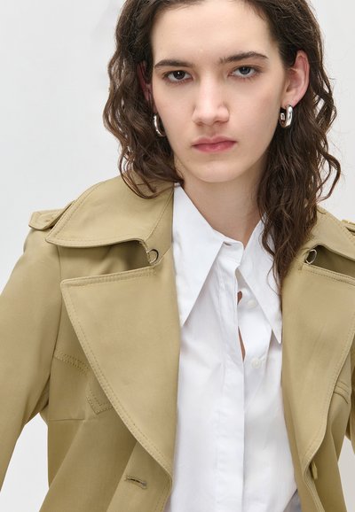 Νεαρή γυναίκα με καστανά σγουρά μαλλιά, που φοράει ασημένια κρίκους σκουλαρίκια, λευκό πουκάμισο με γιακά και μπεζ καμπαρντίνα, σε απλό φόντο.