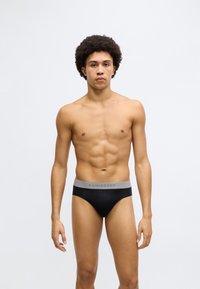 Fekete pamut alsónadrág szürke elasztikus derékpánttal, amelyen a "Schiesser" márkanév olvasható. A modell semleges kifejezéssel áll sima háttér előtt.