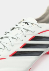 Λευκό ποδοσφαιρικό παπούτσι με μαύρες και κόκκινες λεπτομέρειες. Διαθέτει λείο άνω υλικό, πολλαπλές τρύπες για κορδόνια και υφή κατά μήκος των πλαισίων.