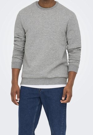 Man in een grijze langemouwen trui over een wit shirt en blauwe jeans, staande met ontspannen armen langs zijn zij.