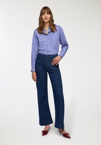 Camicia a righe blu e bianche con bottoni, pantaloni in denim scuro a vita alta, scarpe rosse con tacco e decorazioni a fiocco, modella in piedi con una mano sui fianchi.