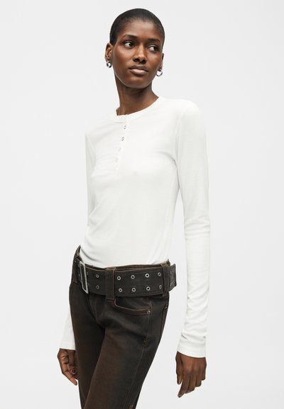 Γυναίκα που φοράει λευκό πουκάμισο με μακριά μανίκια και κουμπιά, μαύρο τζιν και φαρδύ μαύρο ζωνάρι με μεταλλικά χερούλια, κοιτάζοντας προς τα πλάγια.