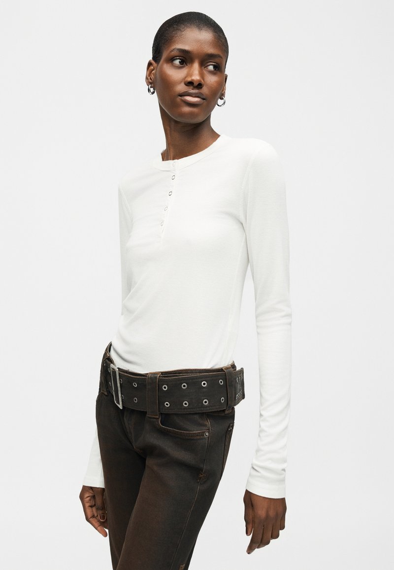 Femme portant une chemise blanche à manches longues boutonnée, un jean noir et une large ceinture noire avec des œillets métalliques, regardant sur le côté.