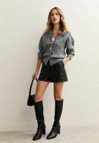 Grijze gebreide cardigan met ruitpatroon, zwarte leren shorts en zwarte kniehoge laarzen met hak. Accessoires bestaan uit een hangerketting en een kleine tas.