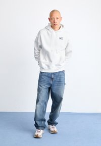 Sweatshirt branco com capuz e bolso frontal, jeans de ganga azuis e ténis bege com atacadores vermelhos. Texturas suaves por todo o lado.