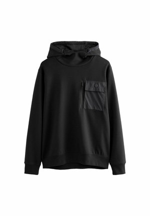 Personne portant un sweat à capuche foncé, un short cargo kaki et des baskets foncées, debout à l'intérieur, devant un mur en panneaux de bois verticaux.