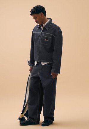 Moški, ki gleda navzdol, nosi temno denim jakno in široke kavbojke, drži črtasto blago na bež ozadju.