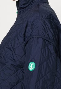 Chaqueta acolchada en color navy con un cuello alto, que presenta un parche de logo circular verde en la manga y una superficie lisa y texturizada.