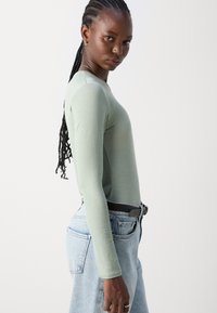 Langermet, lys grønn topp laget av lett materiale, kombinert med høyt liv lys blå denimjeans og et svart belte.