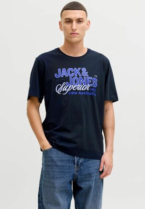 Black cotton t-shirt with blue graphic text "JACK & JONES Superior" on the front, paired with blue denim jeans, worn casually.