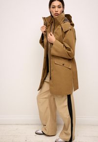 Veste imperméable marron avec un col haut, fermeture à pression sur le devant et poches à rabat. Associée à un pantalon à jambes larges beige avec des bandes noires sur les côtés.