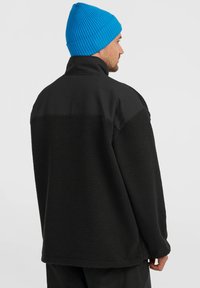 Veste en polaire noire avec un tissu texturé, présentant une partie supérieure noire unie et un bonnet bleu côtelé. Coutures visibles et coupe décontractée.