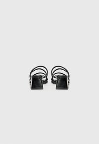 Black open-toed heeled sandals with a square block heel, featuring thin crossed straps across the front and a smooth leather texture.