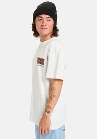 Camiseta blanca de manga corta con un parche de logo cuadrado colorido en el lado izquierdo del pecho y un corte relajado, combinada con un gorro negro.