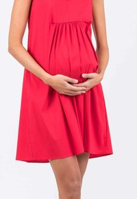 Vestito di cotone rosso con una vestibilità comoda, dettagli arricciati in vita e maniche corte. Mani poggiate su un pancione visibile.