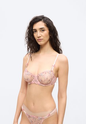 Naine, kellel on pikk tume lokkis juuksed, kannab läbipaistvat roosa lillemustriga tikandiga rinnahoidjat ja sobivaid püksikuid ühevärvilise heleda tausta ees.