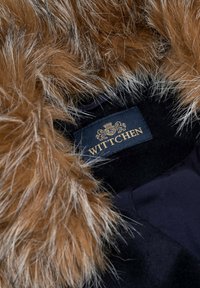 Finitura in pelliccia sintetica marrone su un cappotto di tessuto nero, con un'etichetta che porta il nome "WITTCHEN" in lettere dorate. Aspetto strutturato e lussuoso.