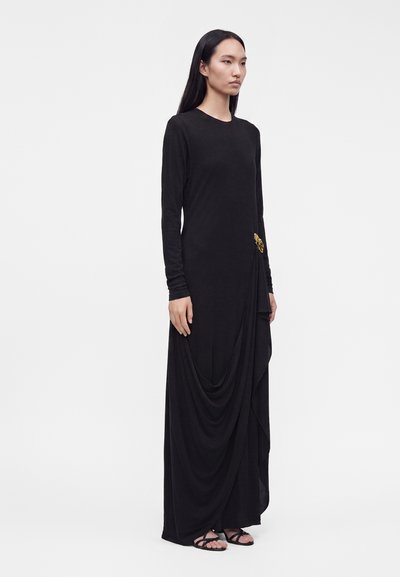 Femme debout dans une longue robe noire à manches longues avec un détail drapé fixé par une broche en or, portant des sandales noires.