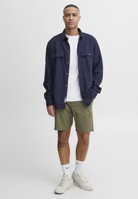 Camisa de botón azul marino con dos bolsillos en el pecho, camiseta blanca, pantalones cortos verdes oliva, zapatillas blancas y un tatuaje texturizado visible en la pierna.