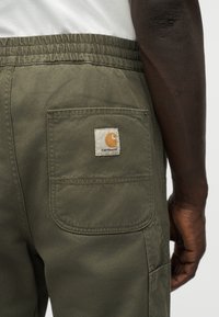 Κοντινό πλάνο σε παντελόνι λαδί με ελαστική μέση και πίσω τσέπη που φέρει το λογότυπο Carhartt σε ένα άτομο κάτω μέρος κορμού και χεριού.