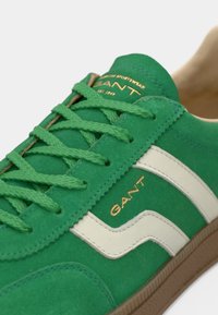 Sneaker in suede verde con dettagli in pelle bianca, lacci testurizzati e marchio dorato. Presenta una suola in gomma e una punta arrotondata.