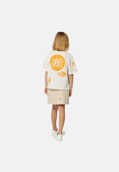 T-shirt blanc surdimensionné avec impression graphique orange, associé à une jupe rayée beige et des baskets blanches. Vue de dos, montrant les détails du design.
