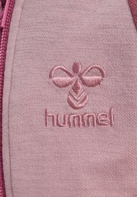 Ljust rosa tyg med en broderad logotyp av en bi och ordet "hummel" nedanför, med en texturerad yta och slät finish.