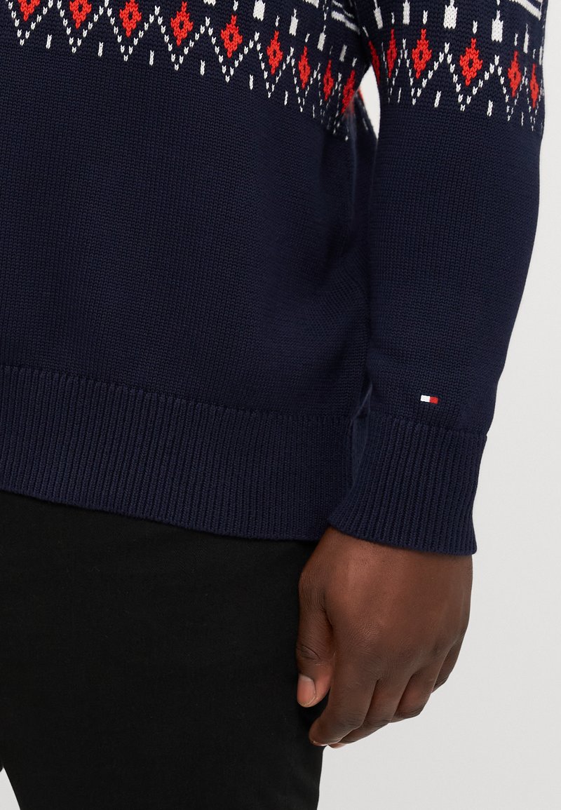 Navy blue knitted sweater featuring a geometric pattern in red, white, and black along the shoulders and sleeves. Ribbed cuffs and hem.