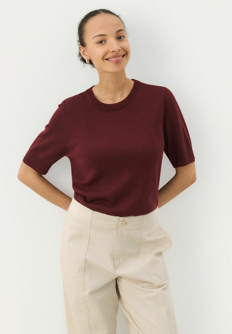Maglione bordeaux a maniche corte in un morbido tessuto a maglia con scollatura rotonda, abbinato a pantaloni beige a vita alta, in piedi contro uno sfondo bianco.