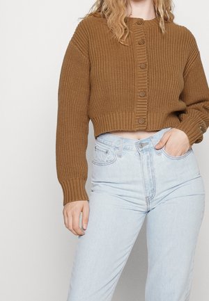 Kobieta ubrana w brązowy krótki sweter z guzikami i jasnoniebieskie, wysoki stan dżinsy, z jedną ręką w kieszeni, na jednolitym tle.