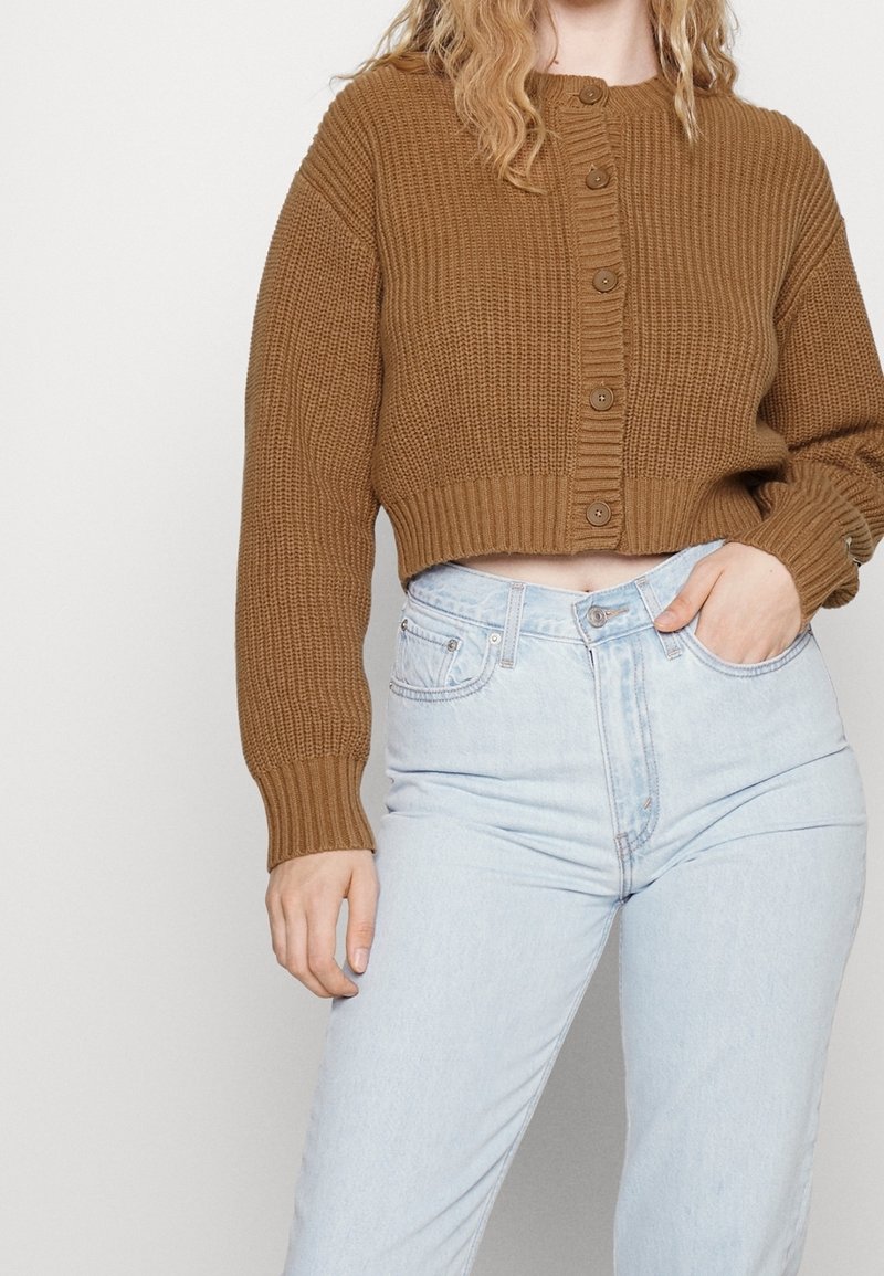 Femme portant un cardigan tricoté marron court à boutons et un jean taille haute bleu clair, une main dans la poche, sur un fond uni.