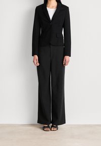 Zwarte getailleerde blazer met een enkele knoop, twee voorzakken, gecombineerd met uitlopende zwarte broeken en platte zwarte sandalen.