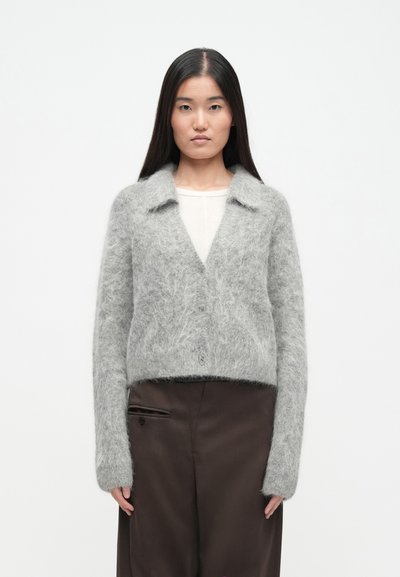 Grijze, pluizige cardigan met een ingesneden kraag, knopen aan de voorkant en lange mouwen; gecombineerd met een bruine broek, voor een gelaagde uitstraling.