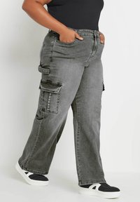 Pantalones cargo en denim gris oscuro con grandes bolsillos laterales, un diseño de pierna recta y detalles de costura. Combinados con zapatillas negras.