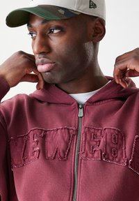 Bordo zip-up kapuca s potrošenim natpisom "NEW" sprijeda, izrađena od mekog materijala. Model nosi kamuflažni šešir.