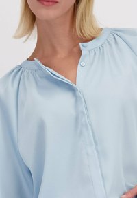 Lichtblauwe blouse met een knoopsluiting bij de hals, geplooide schoudernaden en een gladde, gestructureerde stof. Heeft een relaxte pasvorm en een klassiek ontwerp.