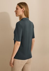 Pánská polokošile s krátkým rukávem z tmavě modré látky, s plochým límcem a hladkým povrchem, nošená s světlými béžovými kalhotami, pohled zezadu.