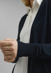 Dunkelblauer Cardigan mit gerippter Textur, offenem Vorderteil und enganliegenden Bündchen. Wird über einem weißen Hemd mit Kragen getragen.