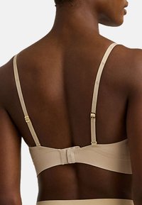 Beige bh med justerbara axelband och hakspänne på baksidan. Tyget är mjukt, med subtil branding på axelbanden.