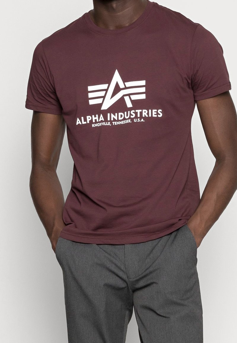 Marošové tričko s krátkým rukávem a bílým logem "Alpha Industries" a geometrickým designem; nošeno s šedými kalhotami, ukazující uvolněný střih.