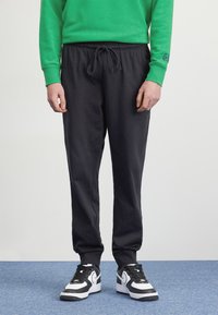 Calças de fato de treino pretas com um cós elástico e cordão, combinadas com sapatilhas desportivas em preto e branco, colocadas contra um fundo simples.