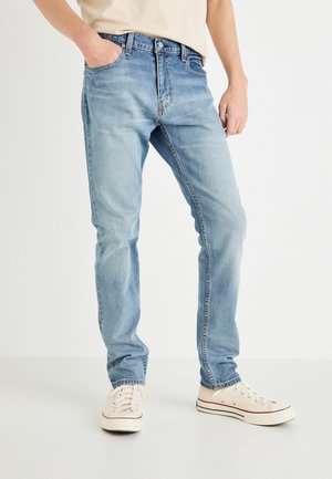 Osoba nosząca jasnoniebieskie dżinsy i białe płócienne trampki stoi na białym tle, z jedną ręką w kieszeni.